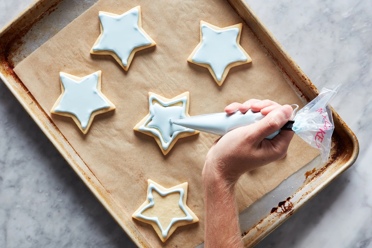 Jak Zrobić Lukier w 5 Minut? Sprawdzona Metoda na Błyszczące Wypieki Hand decorating star-shaped sugar cookies with light blue royal icing on a parchment-lined baking sheet.