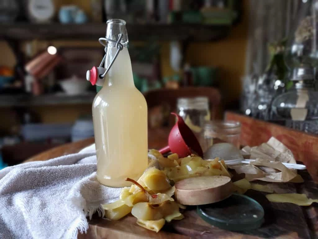 Jak Zrobić Ocet Jabłkowy: Sprawdzony Przepis Z 1932 Roku A glass bottle of homemade apple cider vinegar surrounded by apple scraps and kitchen tools on a wooden surface.