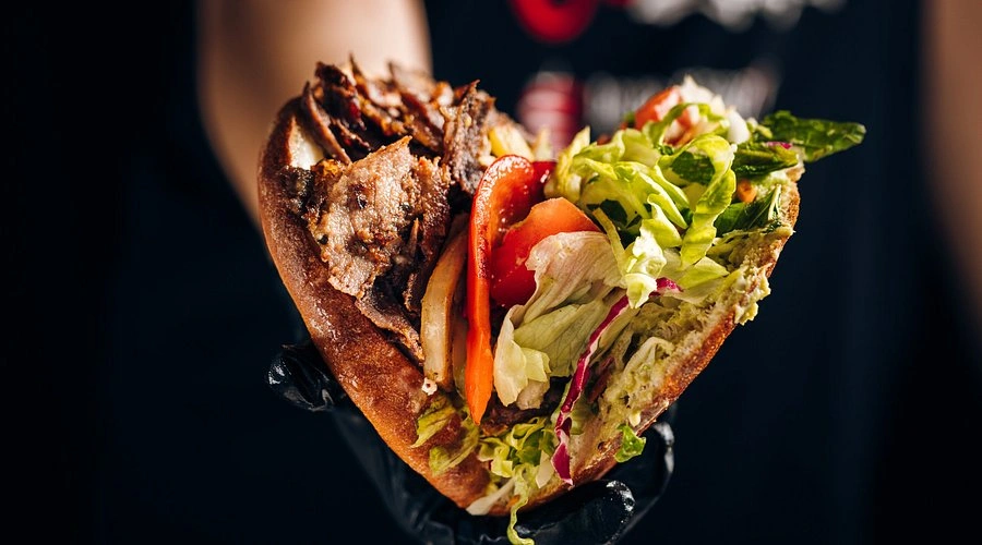 8 Najlepsze Kebaby w Warszawie - Gdzie Zjeść Prawdziwy Turecki Kebab ? Close-up of a hand holding a pita kebab filled with grilled meat, lettuce, tomato, and onions against a dark background.