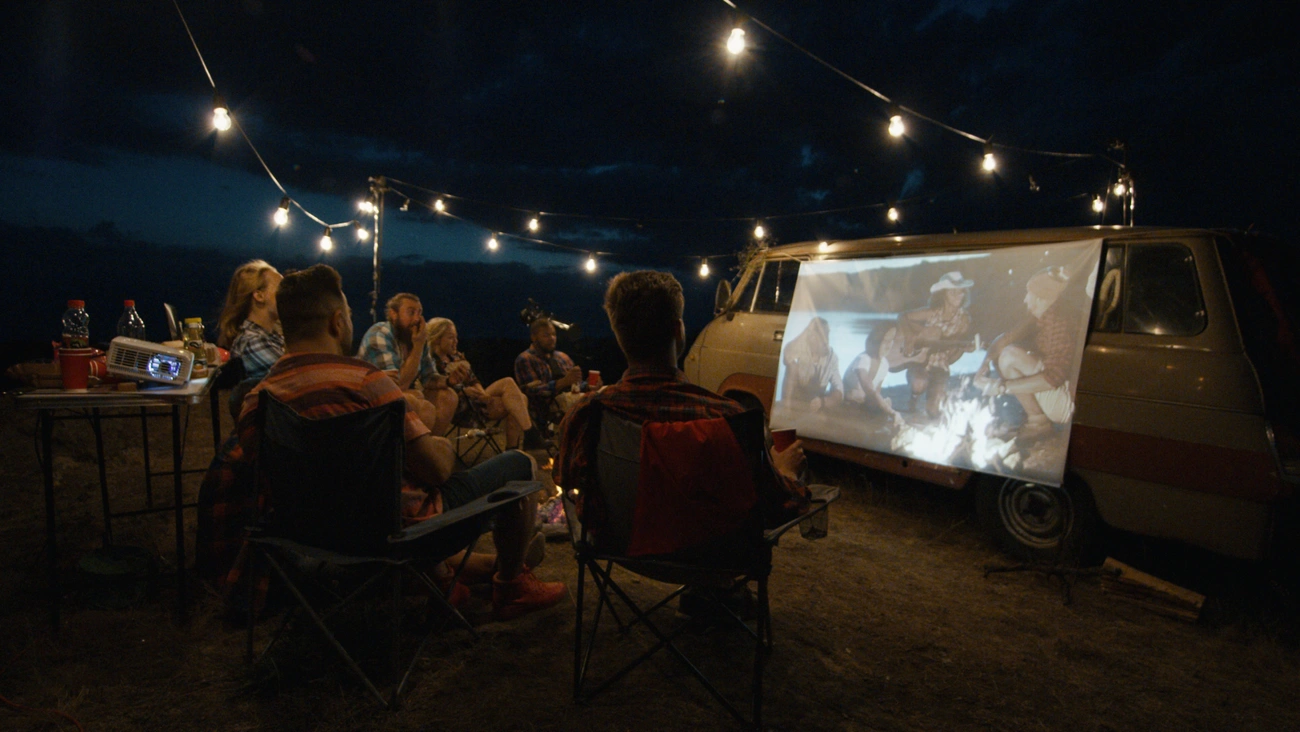 Friends watching an outdoor movie projected on a van screen under string lights on a summer night.