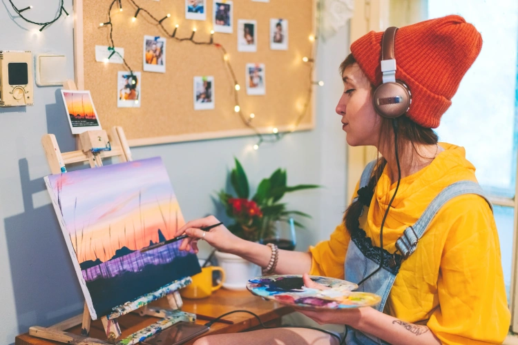 Person wearing a red beanie and headphones painting a sunset landscape on a canvas at home.