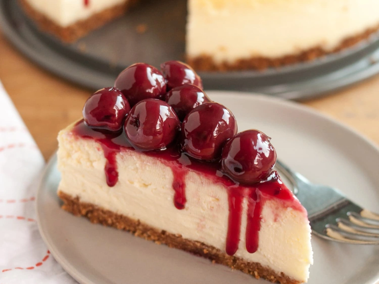 Jak Zrobić Sernik Idealny? Sprawdzone Triki, Które Działają Za Każdym Razem Slice of creamy cheesecake topped with glossy cherry sauce and whole cherries on a plate with a fork.