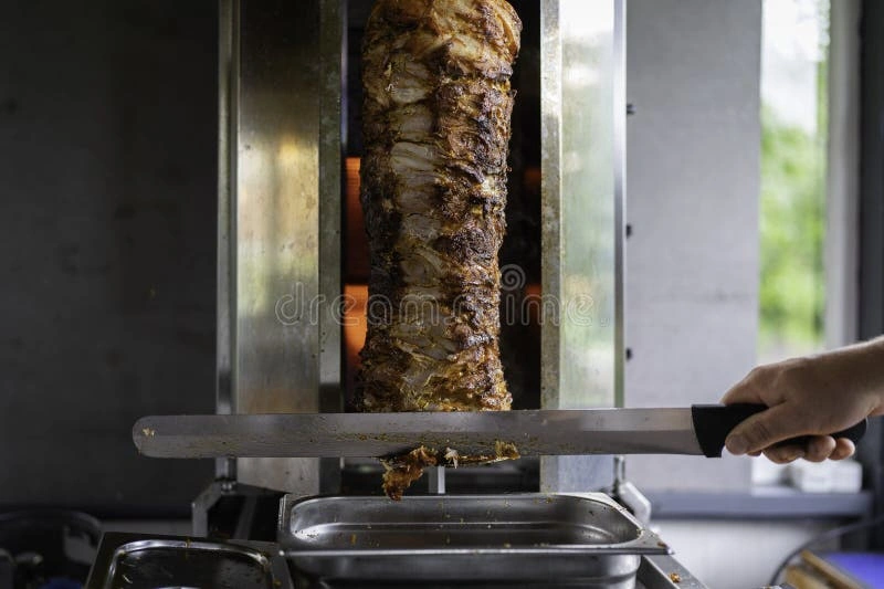 8 Najlepsze Kebaby w Warszawie - Gdzie Zjeść Prawdziwy Turecki Kebab ? Chef slicing freshly cooked traditional Turkish doner kebab meat from a vertical rotisserie in a kitchen setting.
