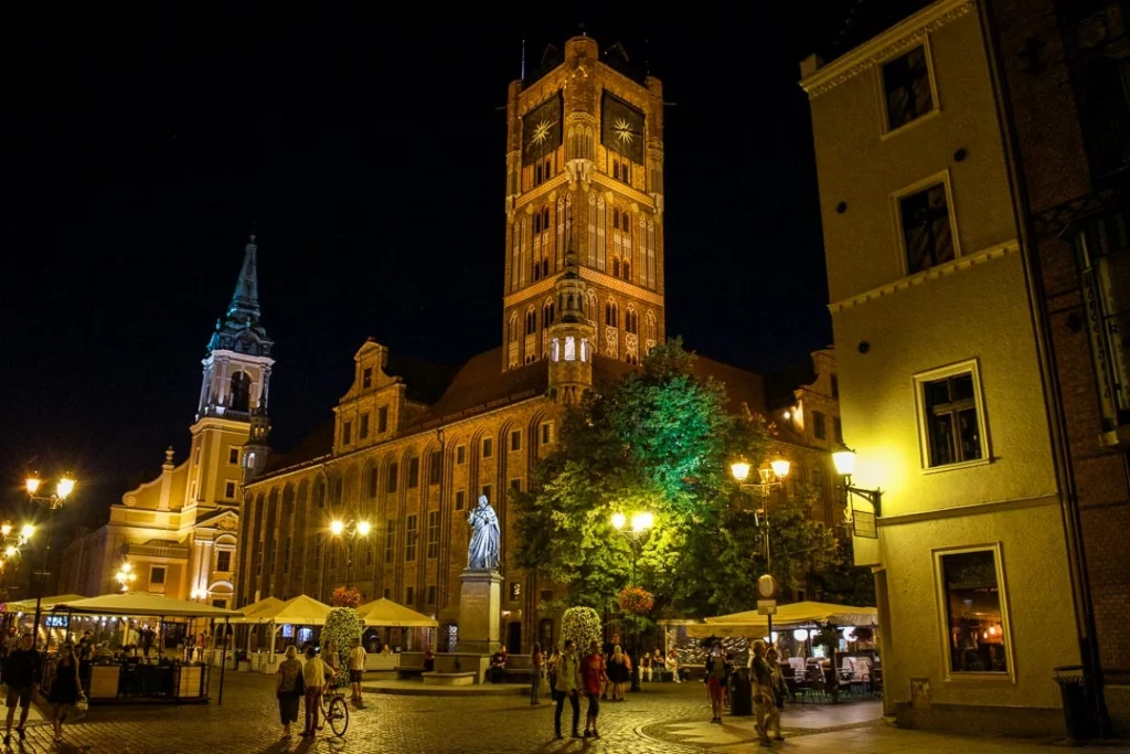 Night view of Torun Old Town in Poland with illuminated historic buildings, a statue, and people walking in the square.