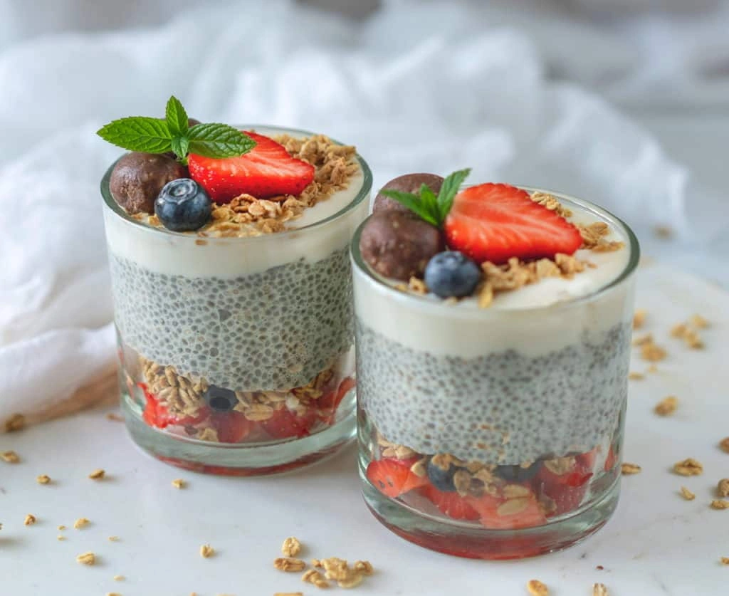 Jak Zrobić Domowe Przekąski Bezglutenowe w 15 Minut Two glasses of vegan chia pudding topped with strawberries, blueberries, granola, chocolate, and mint leaves on a marble surface.