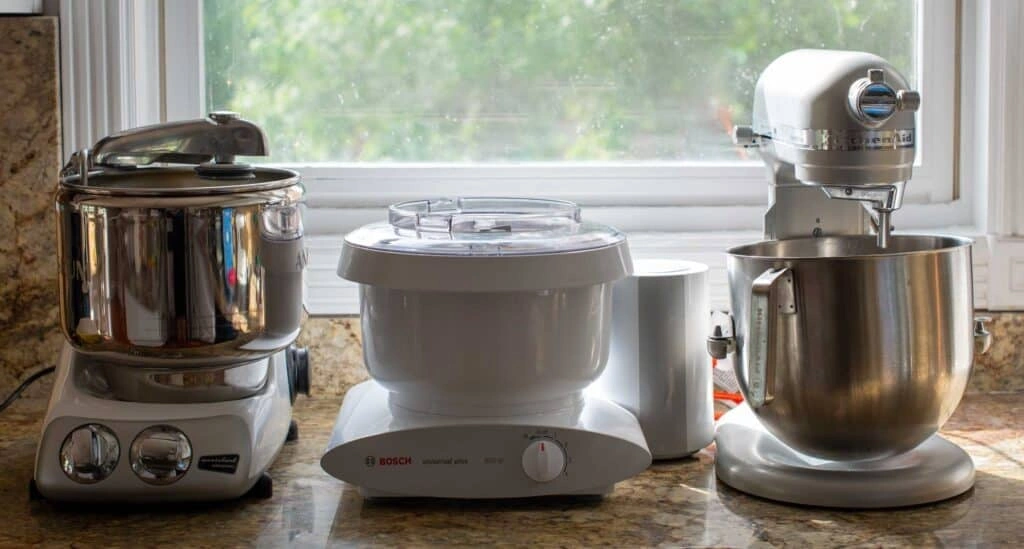 Jak Wybrać Najlepszy Robot Planetarny? Ranking Top 10 + Przewodnik Three stand mixers by Ankarsrum, Bosch, and KitchenAid displayed on a kitchen counter near a window.