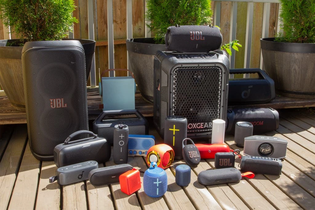 8 Głośników Bluetooth z Najlepszym Dźwiękiem - Ranking 2025 A variety of portable Bluetooth speakers from brands like JBL and Bose displayed outdoors on a wooden deck with plants.