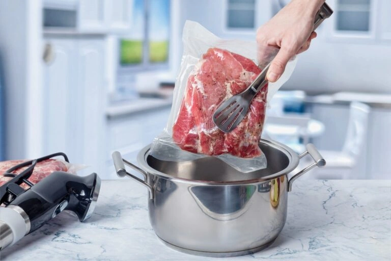 Pierwszy Zakup Sprzętu Sous Vide? Oto Co Musisz Wiedzieć Hand placing vacuum-sealed raw meat into a pot for sous vide cooking in a modern kitchen setting.
