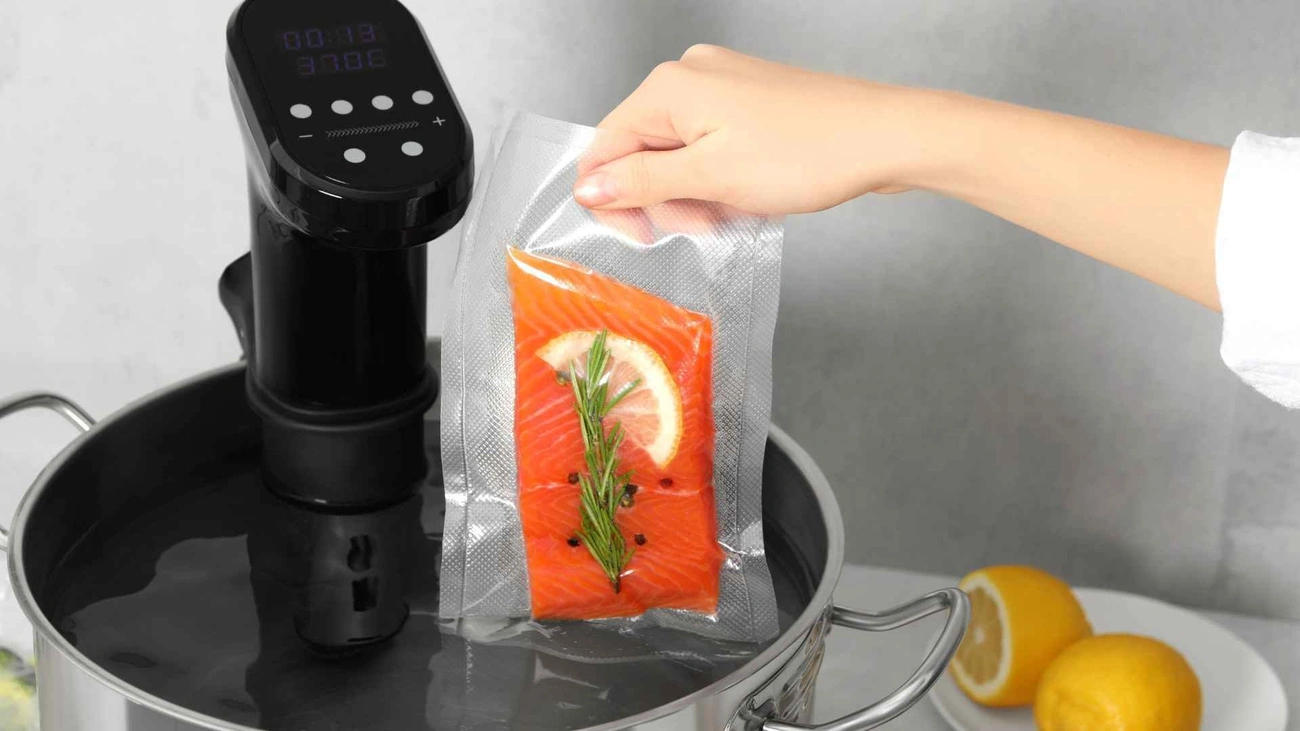 Pierwszy Zakup Sprzętu Sous Vide? Oto Co Musisz Wiedzieć Hand holding vacuum-sealed salmon with herbs next to a sous vide cooker in a water bath pot.