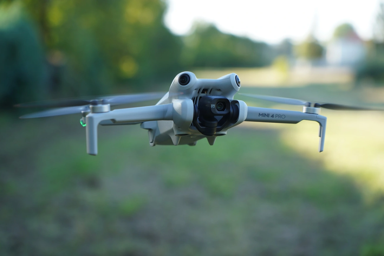 jaki dron na początek? Najlepszy Dron z kamerą: Ranking Top 10 DJI Mini 4 Pro drone hovering outdoors with camera and propellers visible against a blurred background.