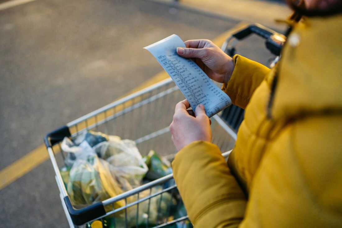 Person in yellow jacket reviews grocery receipt while standing next to a shopping cart filled with produce bags.