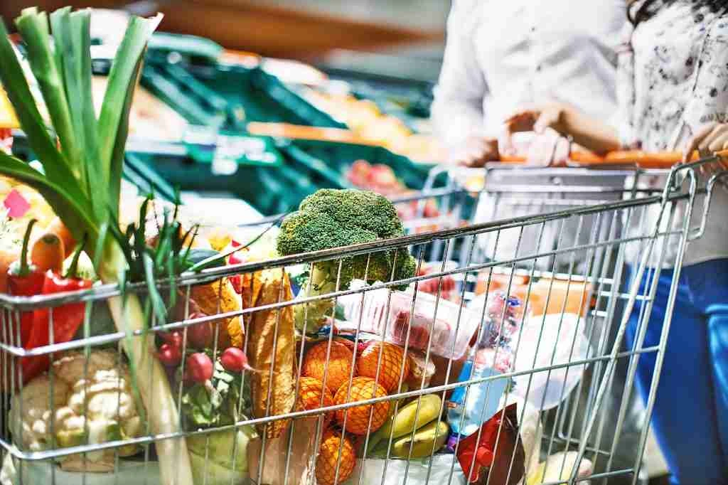 Grocery cart filled with fresh vegetables, fruits, and bread being pushed by shoppers in a supermarket aisle.