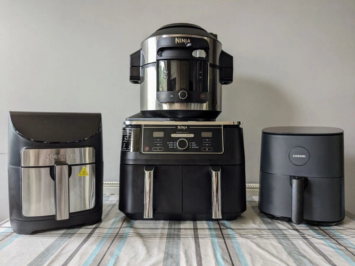 Three family-sized air fryer models from Gourmia, Ninja, and Cosori displayed on a table against a plain wall.