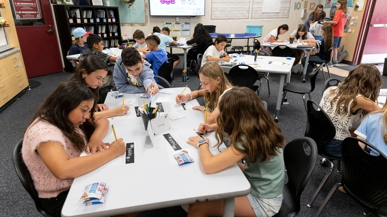 Students in a classroom actively writing at tables with school supplies and snacks visible.