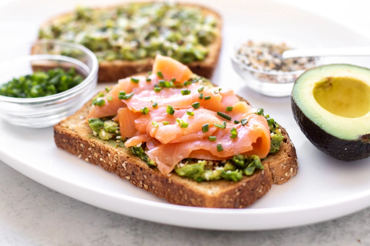 10 Zdrowych Przekąsek do Pracy, Które Dodają Energii na Cały Dzień Smoked salmon slices on avocado spread with chopped chives on multigrain toast served on a white plate with avocado half.