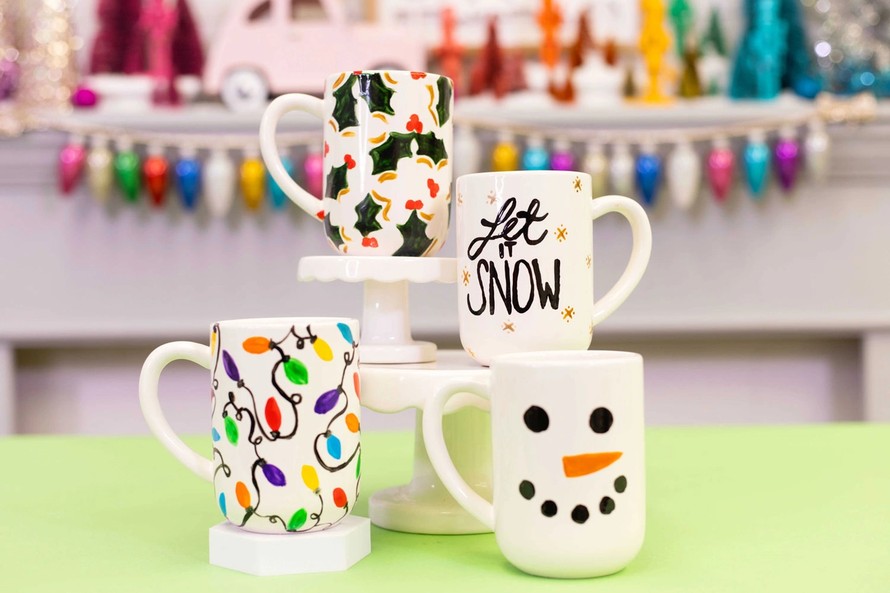 Four hand-painted holiday mugs featuring holly, 'Let it Snow' text, colorful lights, and a snowman face displayed on a green surface.