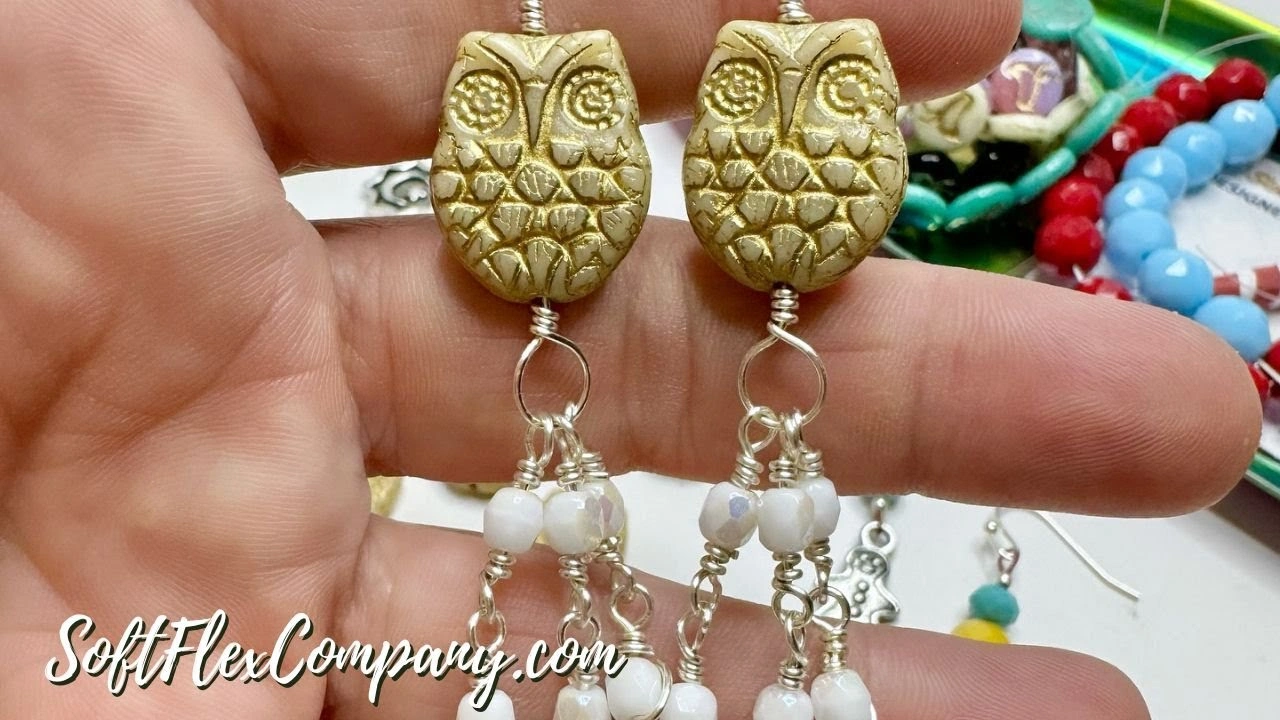 Hand holding owl-shaped gold earrings with dangling white beads, with colorful beaded bracelets in the background.
