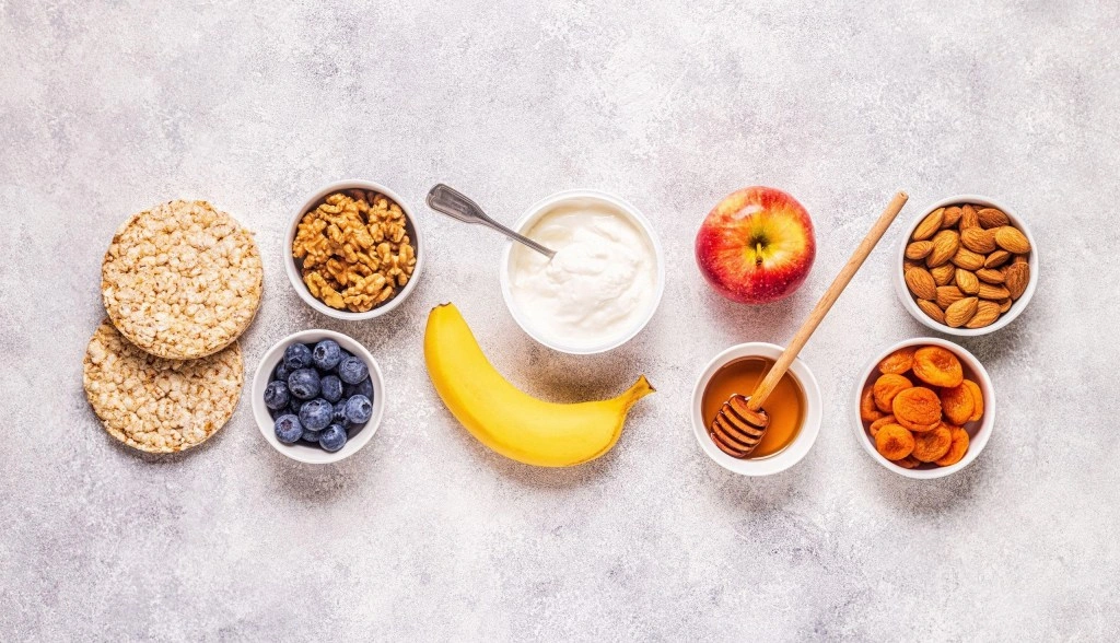 10 Zdrowych Przekąsek do Pracy, Które Dodają Energii na Cały Dzień Assorted healthy office snacks including rice cakes, walnuts, blueberries, banana, yogurt, apple, honey, dried apricots, and almonds on a gray background.