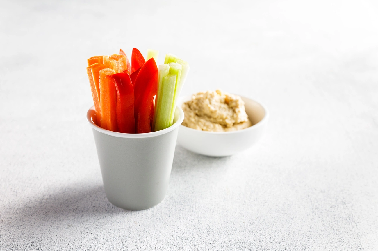 10 Zdrowych Przekąsek do Pracy, Które Dodają Energii na Cały Dzień Cup filled with carrot, red pepper, and celery sticks next to a bowl of hummus on a white surface.