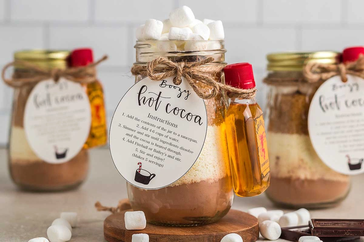 Mason jars filled with layered hot cocoa mix topped with marshmallows and small bottles of liquor attached with twine bows.