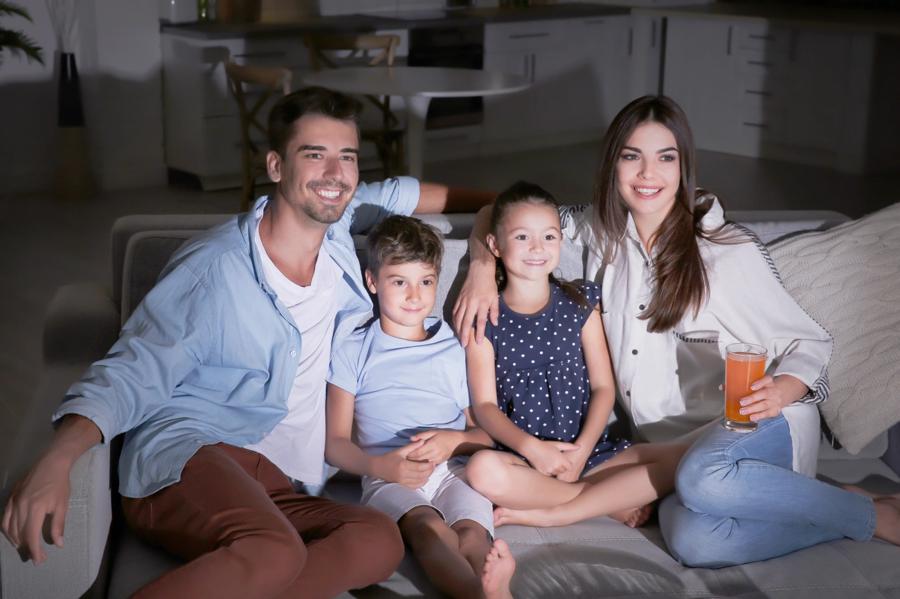 Family sitting together on a couch watching cartoons in a dimly lit living room, with a mother holding a glass of juice.