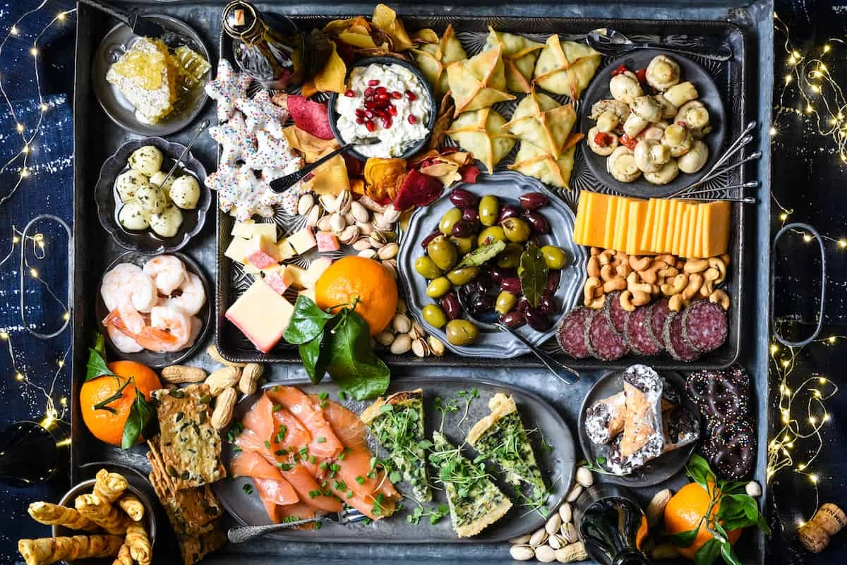 Charcuterie board with assorted cheeses, meats, olives, nuts, smoked salmon, shrimp, dips, and fresh fruit on a dark tray.