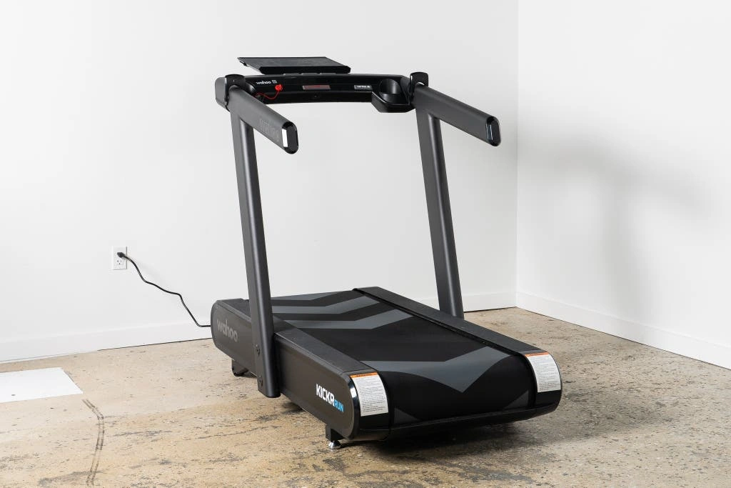Najlepsza bieżnia domowa - Ranking 2026 Black Wahoo KICKR Run treadmill plugged into a wall in a minimalist room with concrete floor and white walls.