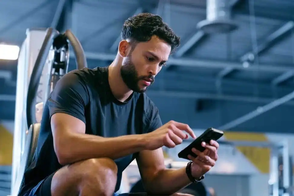 Aplikacje do Ćwiczeń w Domu: Co Naprawdę Działa w 2026? Man in gym wearing black shirt using smartphone while sitting on exercise equipment with blurred background
