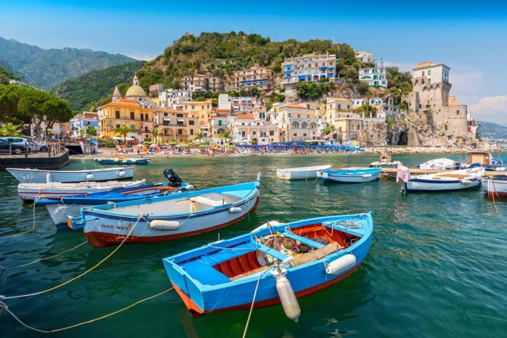 Najlepsze miejsca na wakacje w 2026 roku Colorful boats anchored in the clear water with a scenic Mediterranean hillside village in the background.